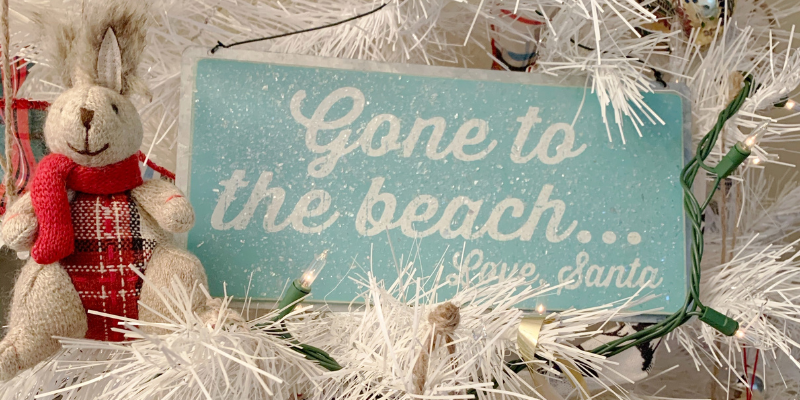 OBX Christmas Ornament - Blue Christmas Ornament that says "Gone to the beach, love Santa" in white font sitting in white Christmas tree. Toy bunny with plaid stomach sitting next to it.