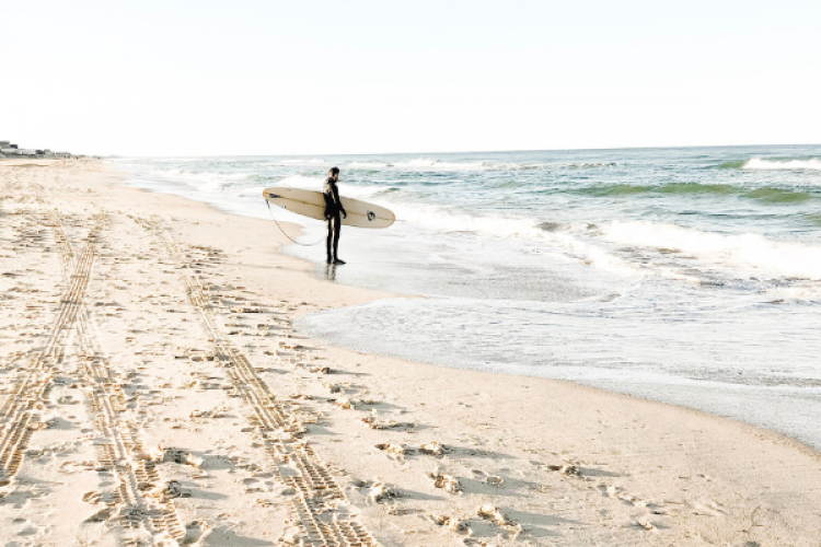 Person standing on shoreline in a wetsuit with a surfboard.