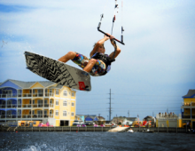 Man in a colorful bathing suit kiteboarding on the sound with a yellow and blue hotel in the background.