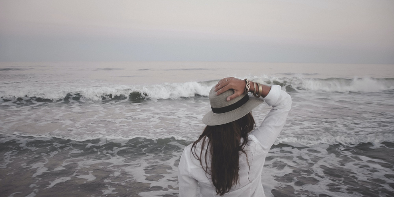 Woman standing in front of gray ocean with hat and sweater.