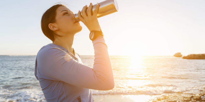 Woman drinking out of silver waterbottle at the beach.