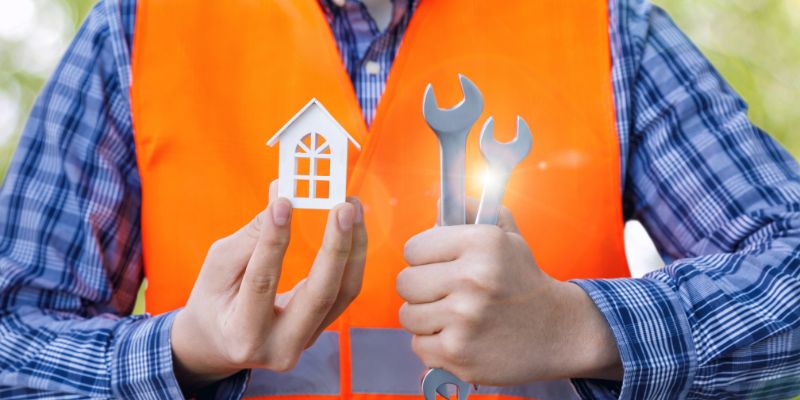 Image of man in an orange vest holding a miniature white house and a wrench.