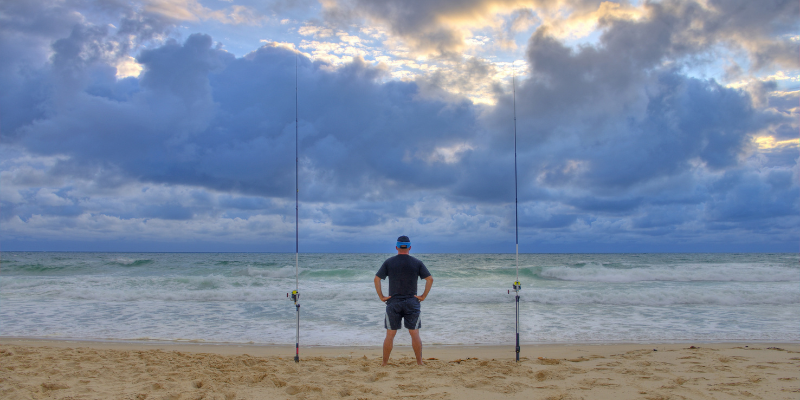 Man standing on the beach with fishing poles in the sand.