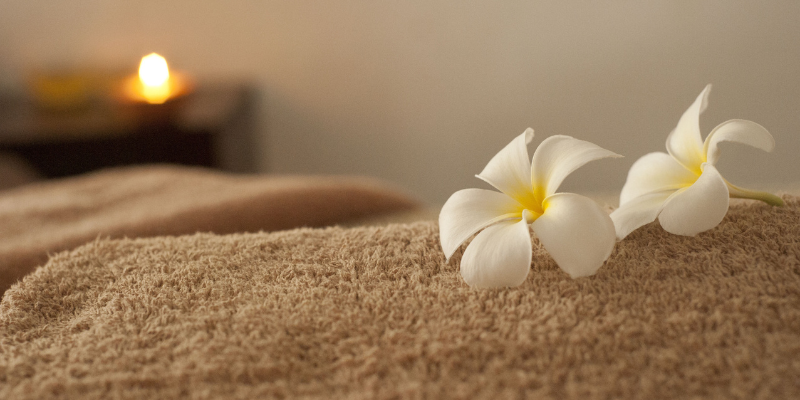 Image of tan towel with white flower sitting on top of it; unfocused image of candle in the background.