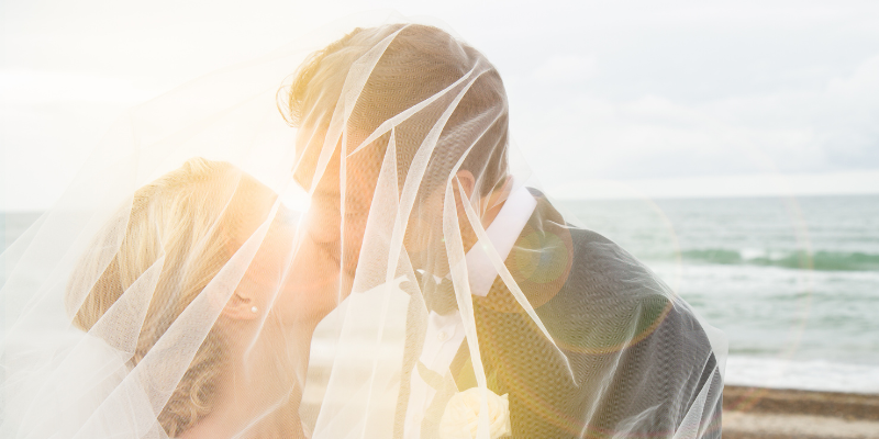 Couple on the beach kissing in front of ocean under wedding veil.
