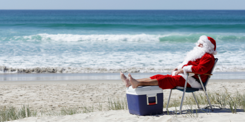 A man dressed as Santa sits on an Outer Banks beach with his feet propped up on a cooler. 