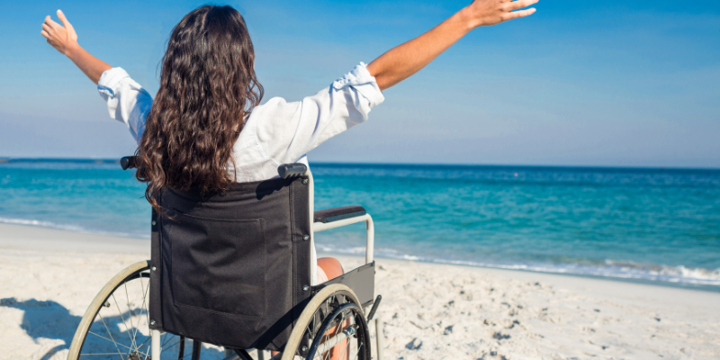 Woman in a wheelchair at the beach with her arms raised to the sky.