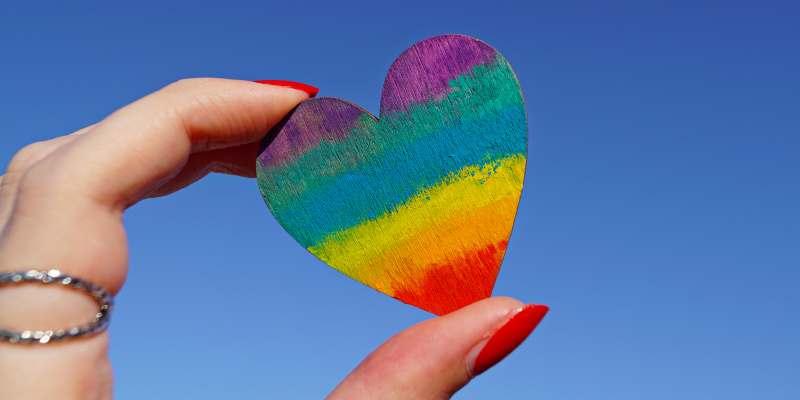 Hand with red fingernails holding a small wooden heart painted rainbow colors.