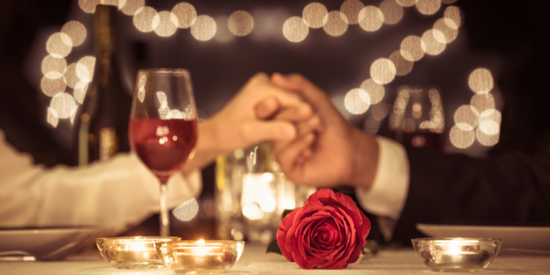 Out of focus image of man and woman holding hands across table; focus on red rose.
