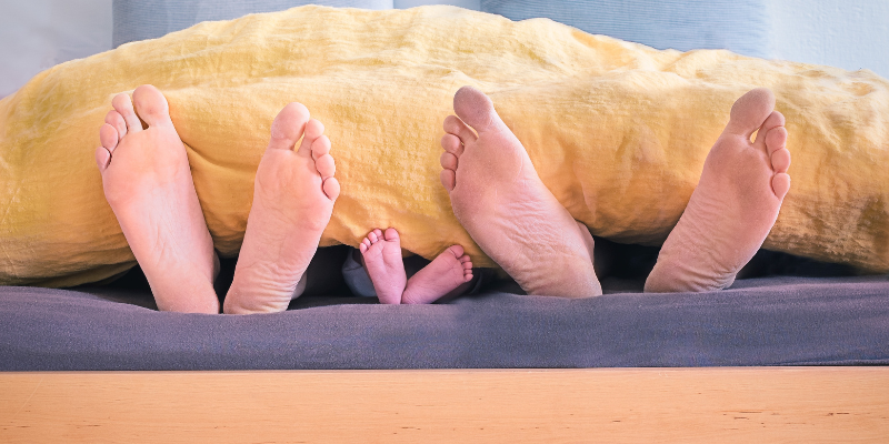 Three sets of feet sticking out from under a blanket; two adult pairs of feet and a baby pair of feet.